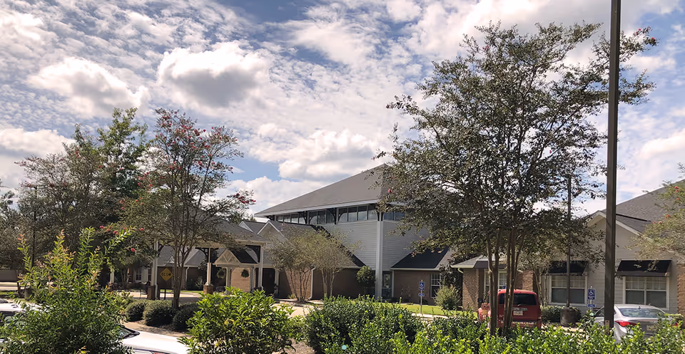 Exterior view of Oak Park Village at Slidell facility showing a large building with multiple rooflines, surrounded by trees and bushes under a partly cloudy sky. Several parked cars and handicap parking signs are visible in the foreground.
