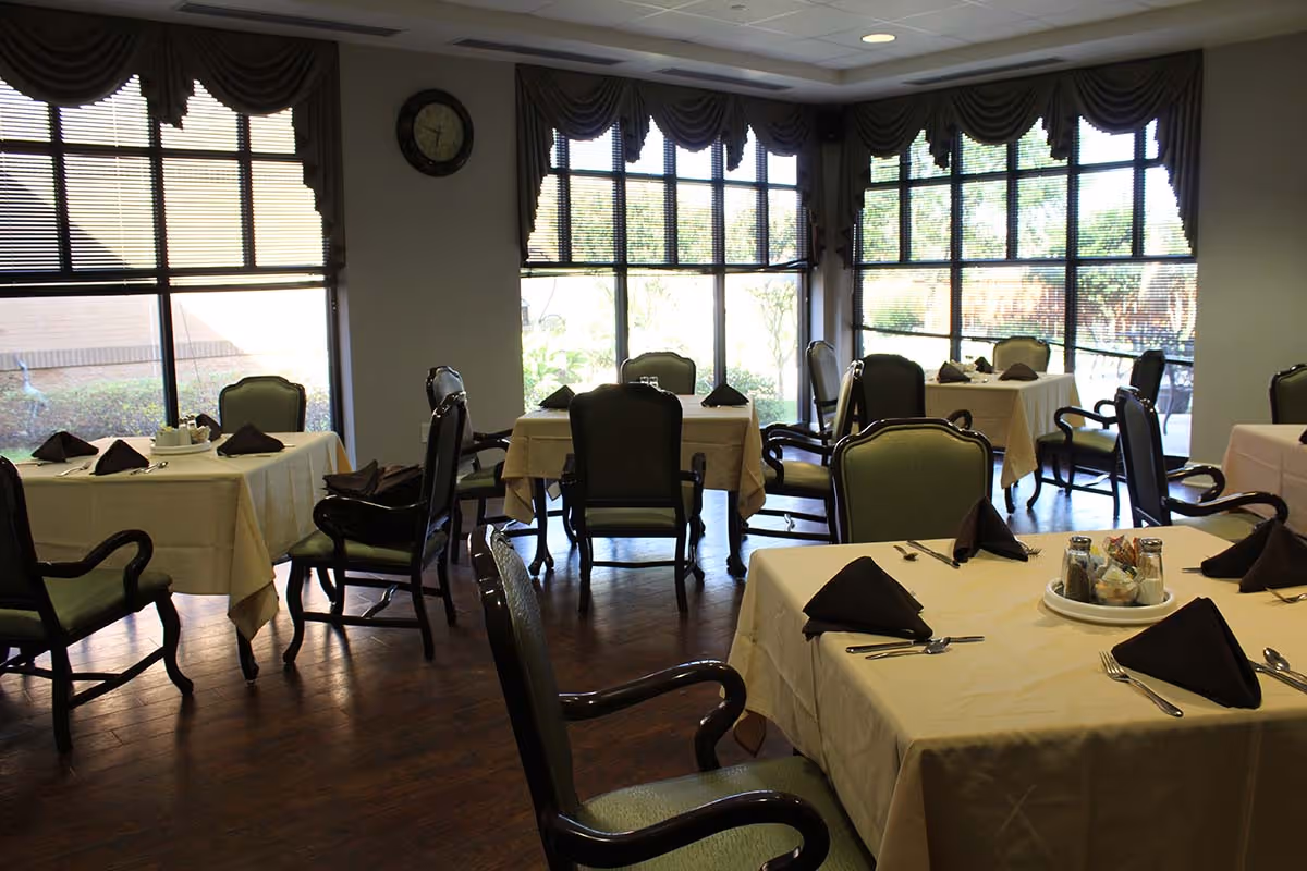 A dining room with several tables covered in beige tablecloths, each set with dark brown folded napkins, silverware, and condiments. The room has large windows with blinds and valances, allowing natural light to brighten the space. Green cushioned chairs with wooden armrests surround the tables, and a clock is mounted on the wall between the windows.