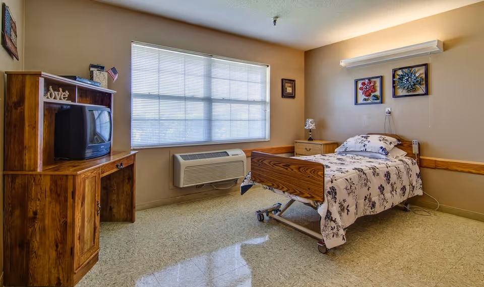A simple, clean bedroom in a senior living facility with a single bed featuring floral bedding, a wooden nightstand with a lamp, a wooden desk with an old-style TV, and a large window with blinds letting in natural light. The walls are beige with two floral wall decorations above the bed.
