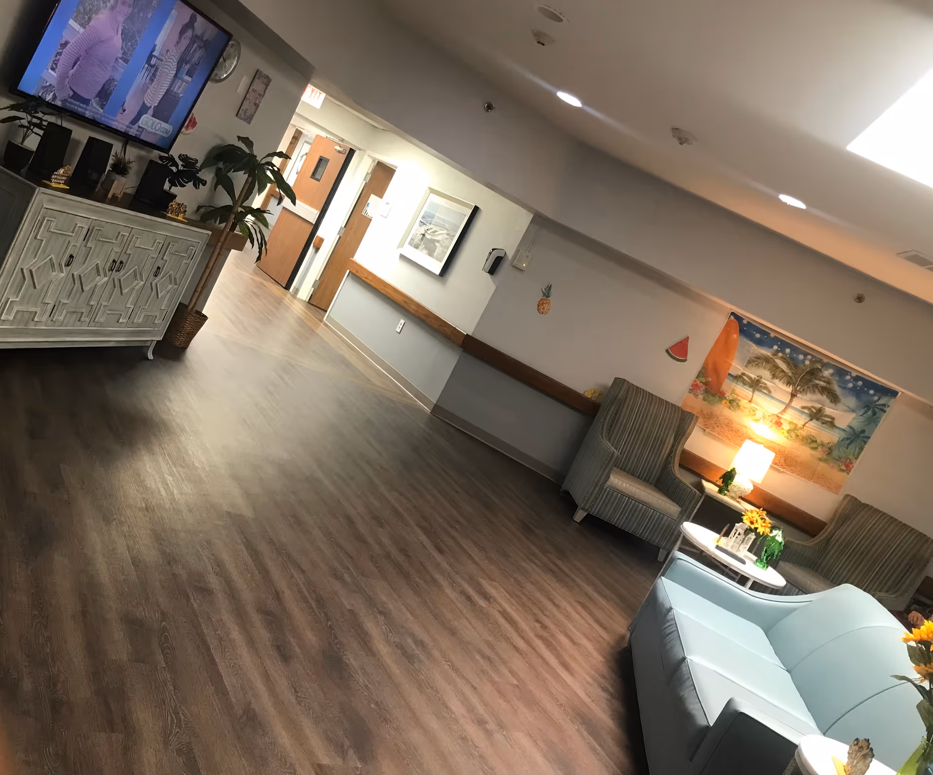 A senior living facility lounge with sofas and armchairs, a TV on a cabinet, wall art and wood-look flooring.