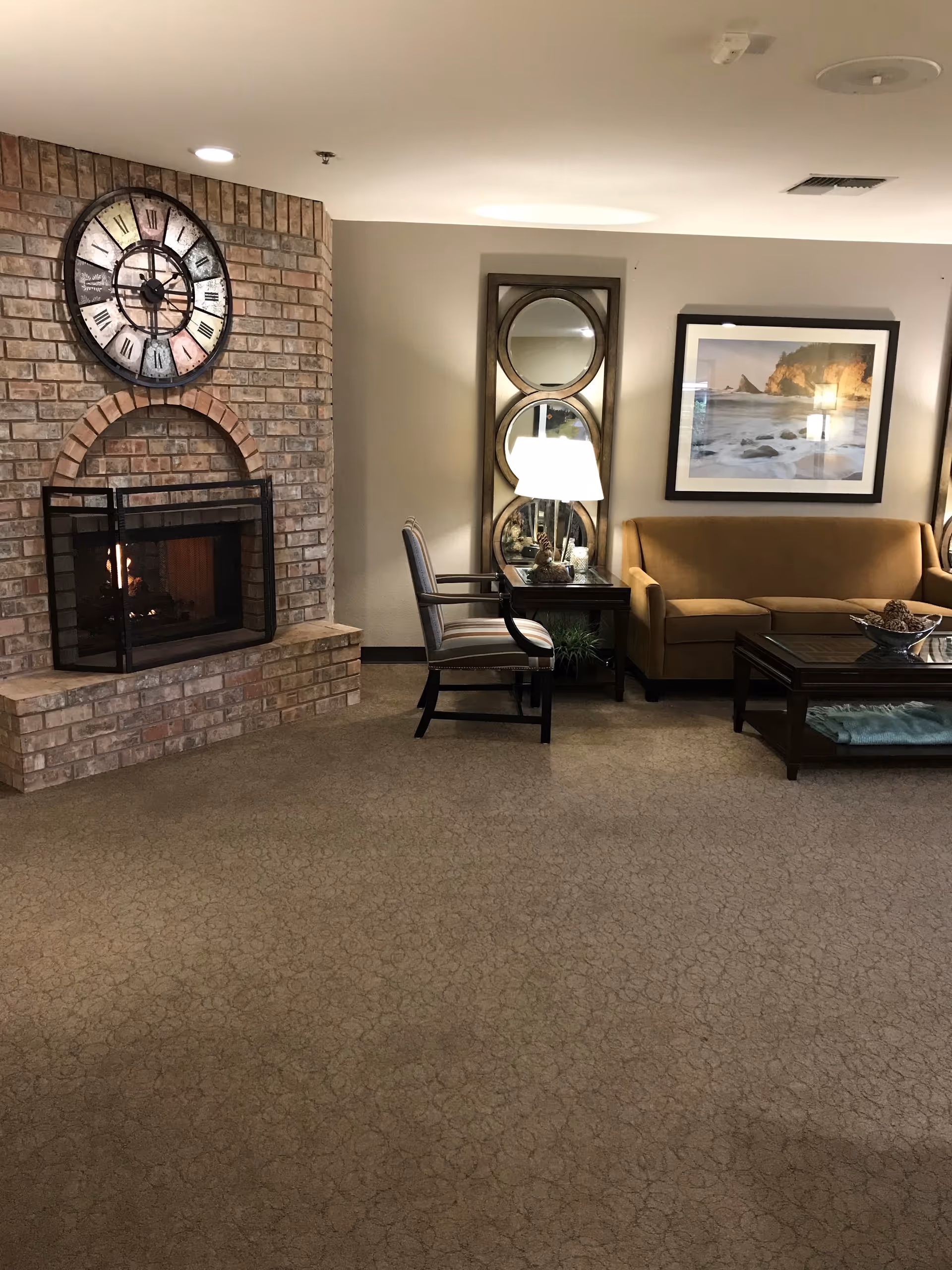 A cozy living room area with a brick fireplace on the left side, a large decorative clock above it, a beige sofa, a wooden coffee table with a decorative bowl, a side table with a lamp, a chair, a tall mirror with three circular sections, and a framed picture of a rocky beach on the wall.