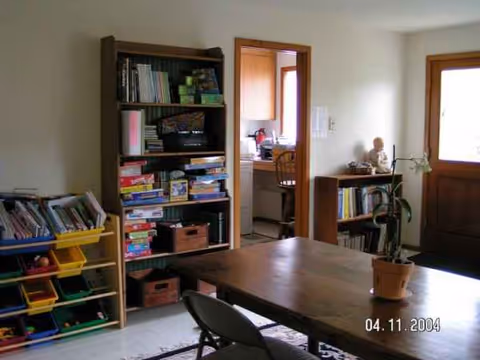 A wood dining table with chairs, bookshelves, and a potted plant in a well-lit room.
