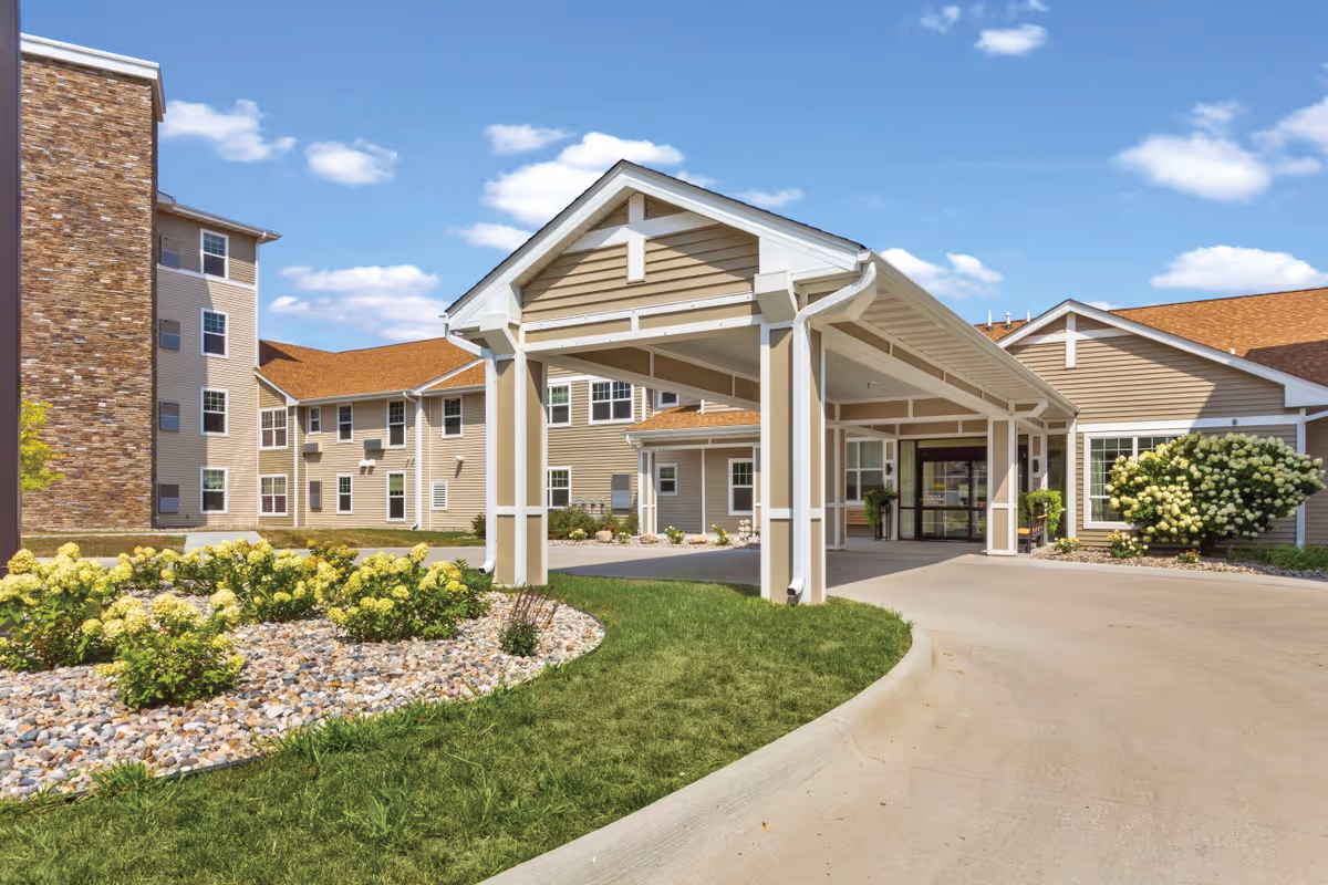 Exterior view of Mill Pond senior living facility showing the main entrance with a covered driveway, beige siding, stone accents, landscaped garden with yellow flowers, and a clear blue sky with some clouds.