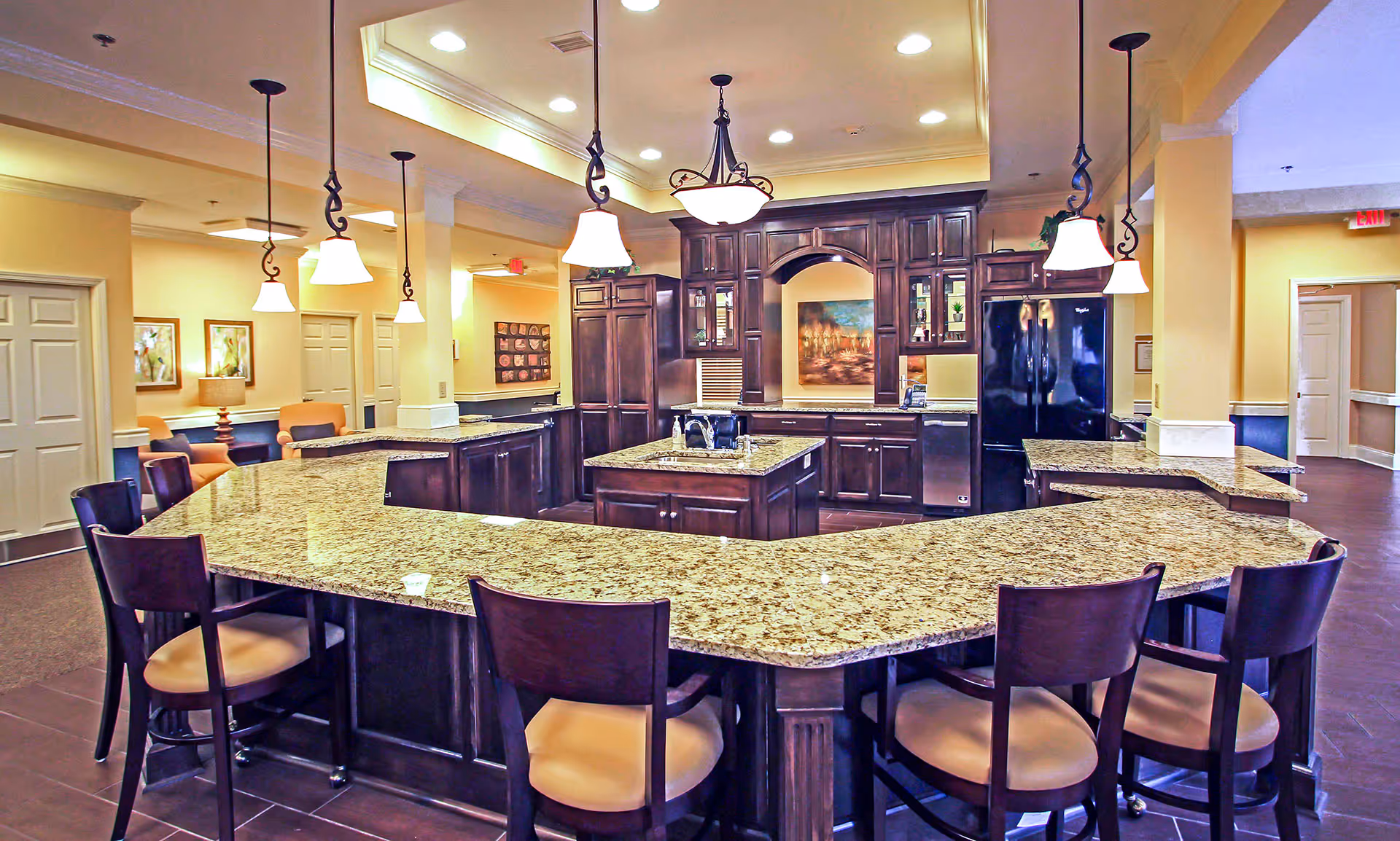 Spacious kitchen area with a large granite countertop island surrounded by wooden chairs with tan cushions. The kitchen features dark wood cabinets, a black refrigerator, a sink on the island, and multiple hanging pendant lights. The background shows a hallway with doors and framed artwork on the walls.