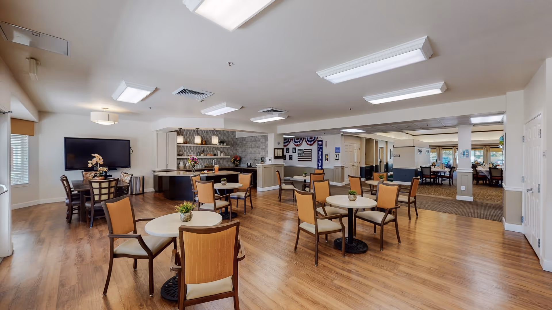A spacious common area in a senior living facility featuring multiple round tables with chairs, a small counter area with pendant lighting, and a large flat-screen TV mounted on the wall. The room has wood flooring, neutral-colored walls, and ceiling lights, with additional seating and dining areas visible in the background.