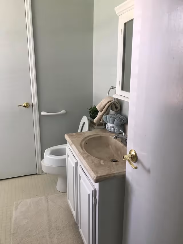 A small bathroom with a white toilet, a beige countertop sink with a cabinet underneath, a white medicine cabinet with a mirror above the sink, and a towel rack holding beige and blue towels. There is a white door with a gold handle and a grab bar on the wall next to the toilet.