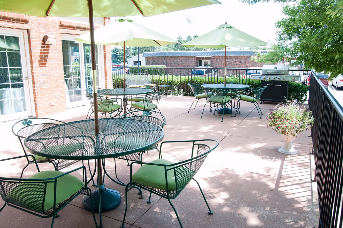 Outdoor patio with green metal tables and chairs, umbrellas, and a grill beside a brick building.
