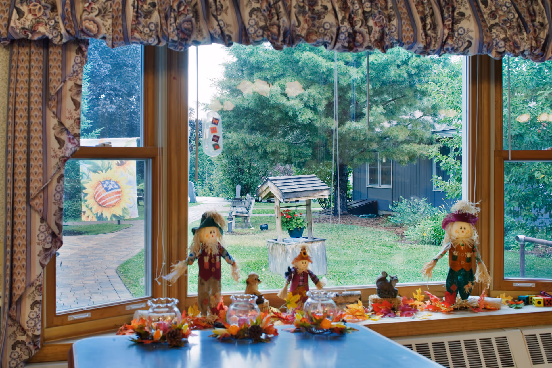 Large window with autumn-themed decorations and scarecrow figurines on a windowsill overlooking a grassy courtyard with a small well and trees.