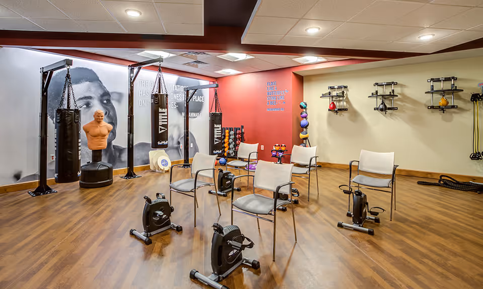 A fitness room with wooden flooring featuring exercise equipment including stationary pedal machines in front of chairs, hanging punching bags, a mannequin torso for boxing practice, and various weights and exercise balls on racks. The walls have motivational quotes and a large black and white mural of a person's face.