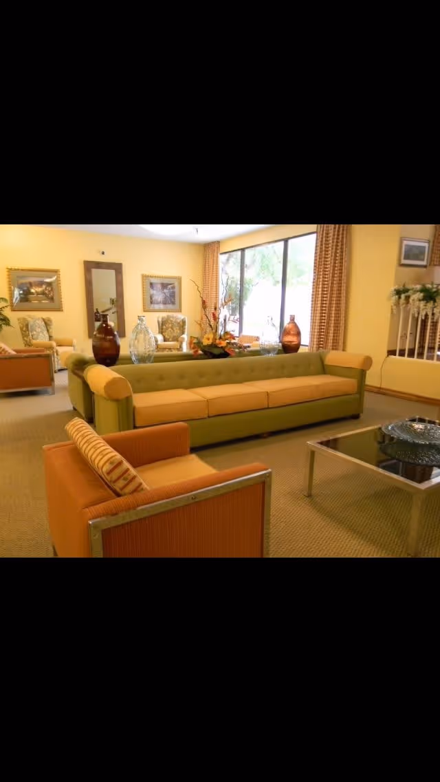 A bright living room area with a long green and beige sofa, an orange armchair with a striped cushion, a glass coffee table, and decorative vases on the sofa. The room has large windows with patterned curtains, framed pictures on the yellow walls, and a mirror. There is also a railing with white flowers on the right side.