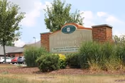 Outdoor view of a brick sign for Golden Years Homestead, Inc. surrounded by landscaping with bushes and grass, with a few parked cars and trees in the background under a partly cloudy sky.