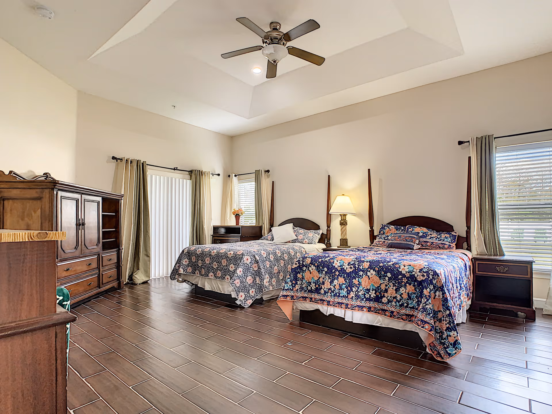 A spacious bedroom with two beds covered in floral bedspreads, a wooden dresser, nightstands, a lamp, and windows with curtains. The room has a ceiling fan and wood-look tile flooring.