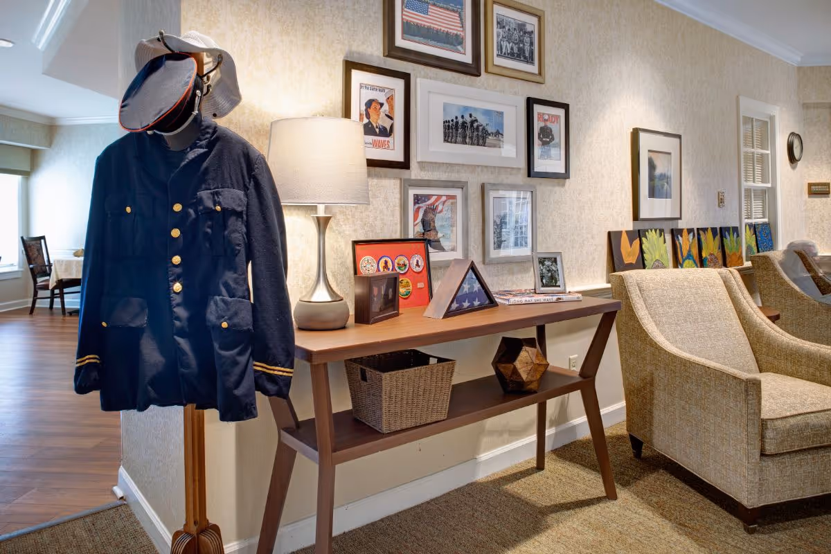 A cozy interior corner of a senior living facility featuring a wooden coat rack with a navy blue military jacket and hats, a wooden console table with a lamp, framed photos, a folded American flag in a display case, and decorative items. Beige upholstered armchairs are positioned nearby, and the wall behind is adorned with various framed pictures and artwork.