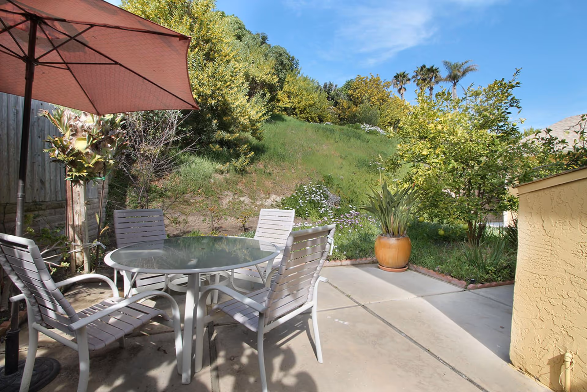 Outdoor patio area with a round glass table and four white chairs under a large red umbrella. The patio is surrounded by greenery including trees, bushes, and a grassy hill under a clear blue sky.