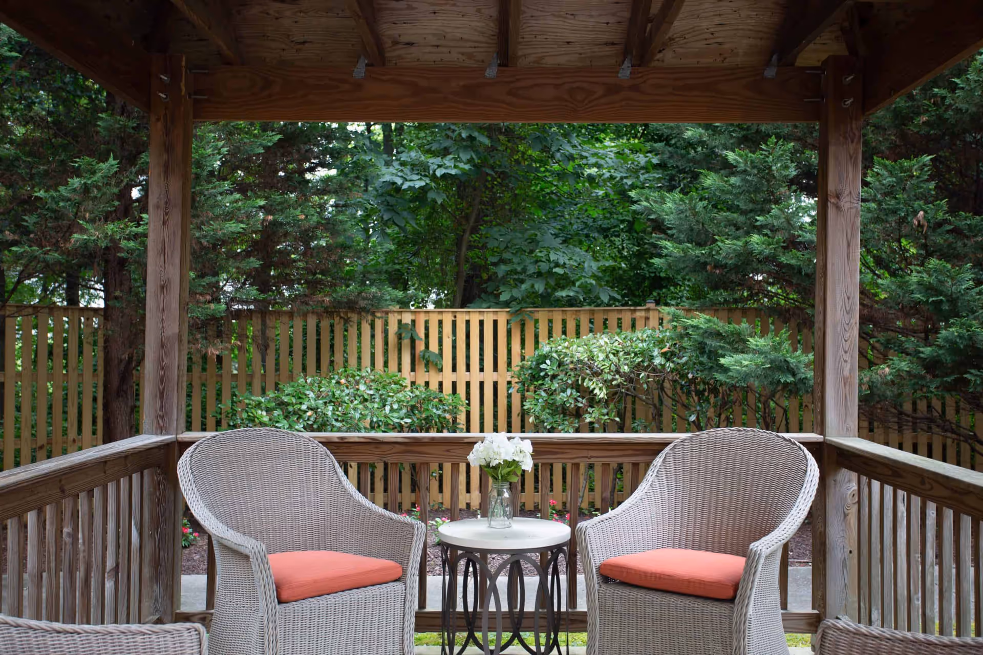 Covered wooden patio with two wicker chairs with orange cushions and a small round table holding a vase of flowers overlooking a fenced garden.