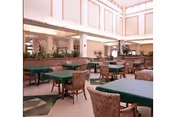 Spacious dining area with multiple tables covered in green tablecloths and surrounded by cushioned chairs. The room features high ceilings with large windows allowing natural light to fill the space, and there are plants placed along the perimeter.