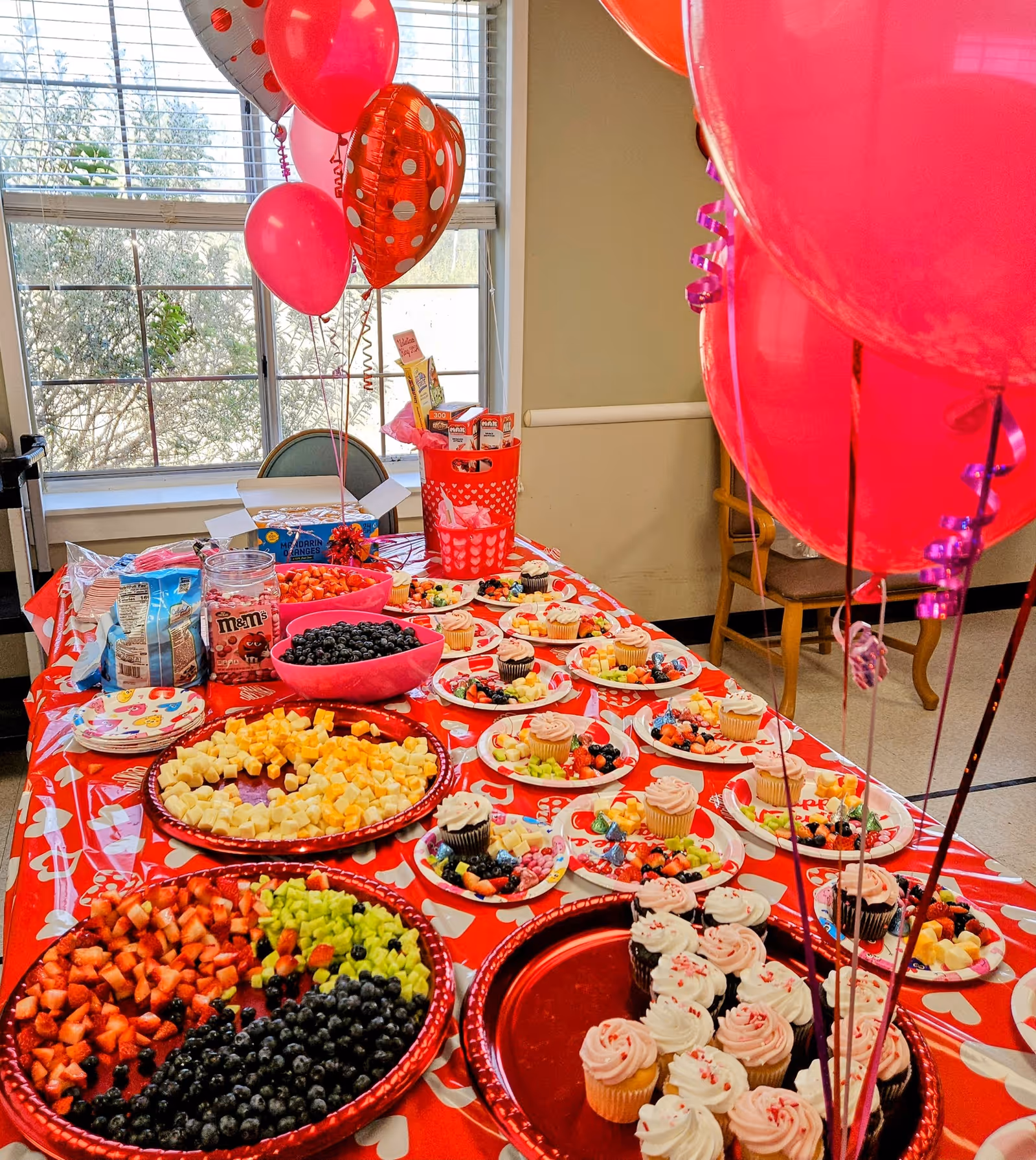 A festive table set up with red and white heart-themed tablecloth and balloons, featuring plates of assorted fresh fruit, cupcakes with pink and white frosting, bowls of blueberries and candy, and various snacks. The setting is indoors near a window with natural light.