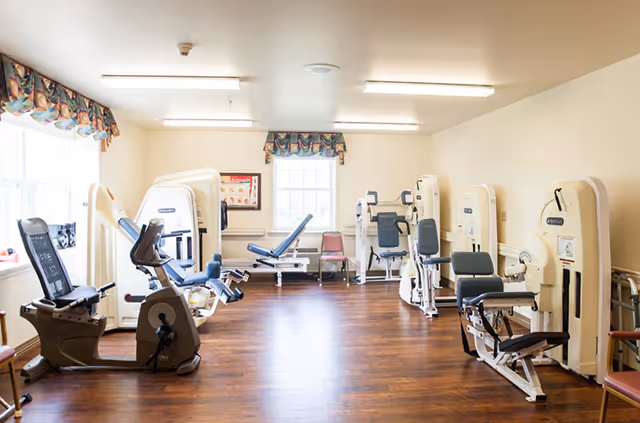 Well-lit fitness room with multiple seated strength machines, an exercise bike, and a weight bench on wood floors near windows.