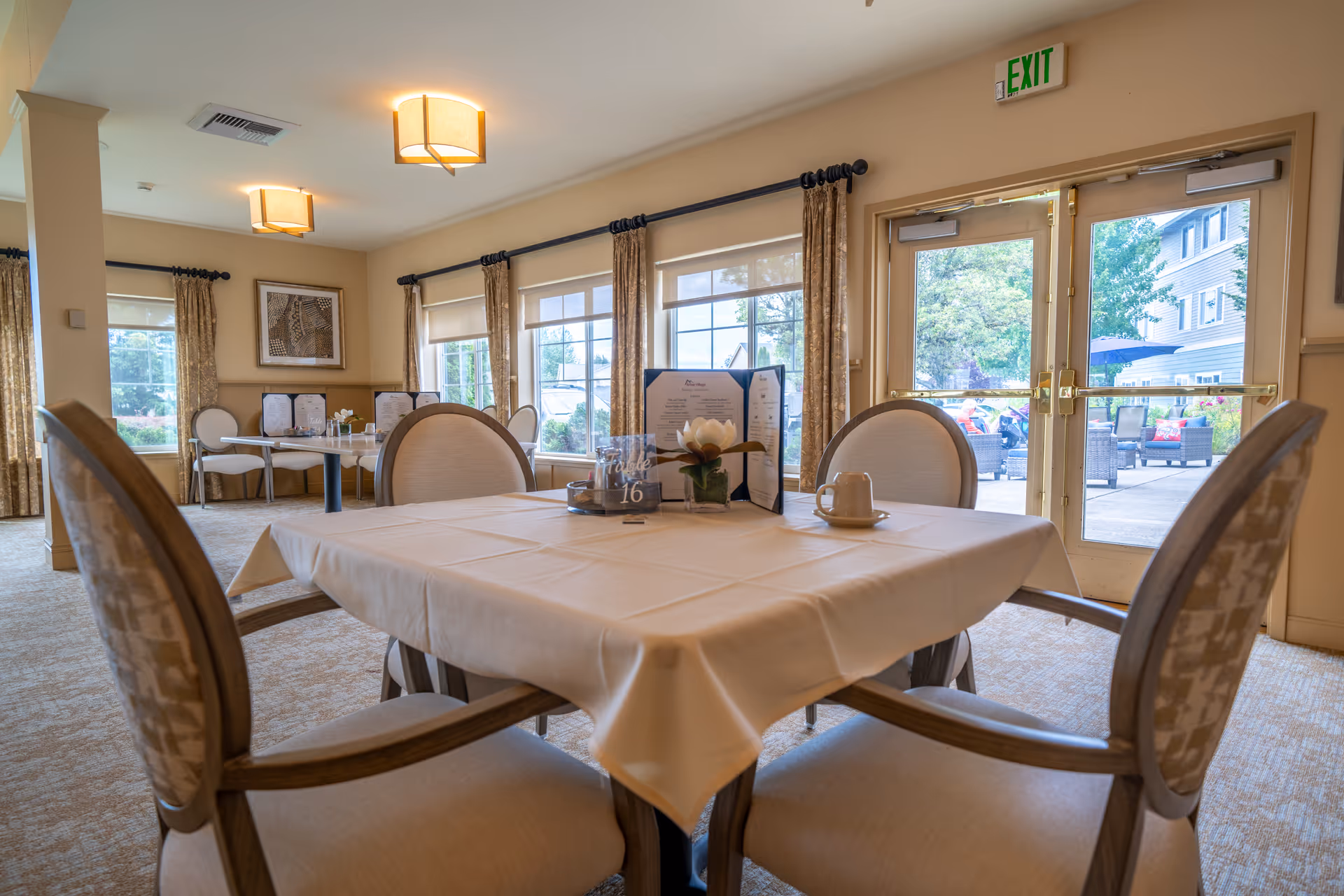 A dining room in a senior living facility with a table covered by a white tablecloth and four cushioned chairs around it. The table has a small flower centerpiece, a menu, and a table number sign. Large windows and glass doors provide a view of an outdoor patio area with seating. The room is softly lit with ceiling lights and decorated with curtains and framed artwork.