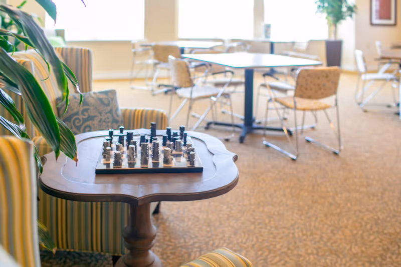 Bright senior living common room with a chessboard on a small wooden table and chairs and dining tables in the background.