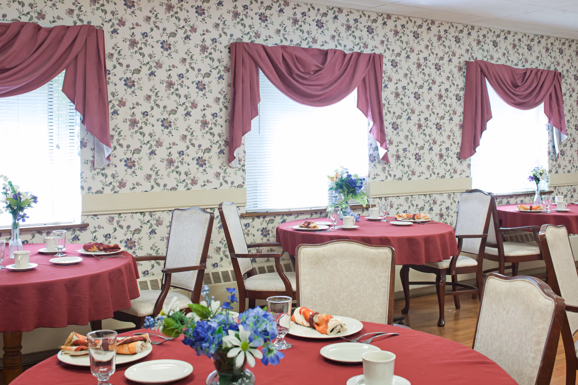 Dining room with round tables covered in red tablecloths, set with plates, cups, glasses, and napkins. The room has floral wallpaper and windows with pink draped curtains. There are upholstered chairs around the tables and floral centerpieces on each table.