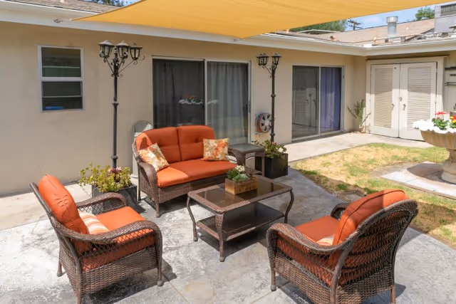 Outdoor patio area with wicker furniture including a loveseat, two armchairs, and a coffee table, all with orange cushions. The patio is shaded by a yellow canopy and surrounded by a beige building with sliding glass doors and windows. There are two black lamp posts and some potted plants around the seating area.