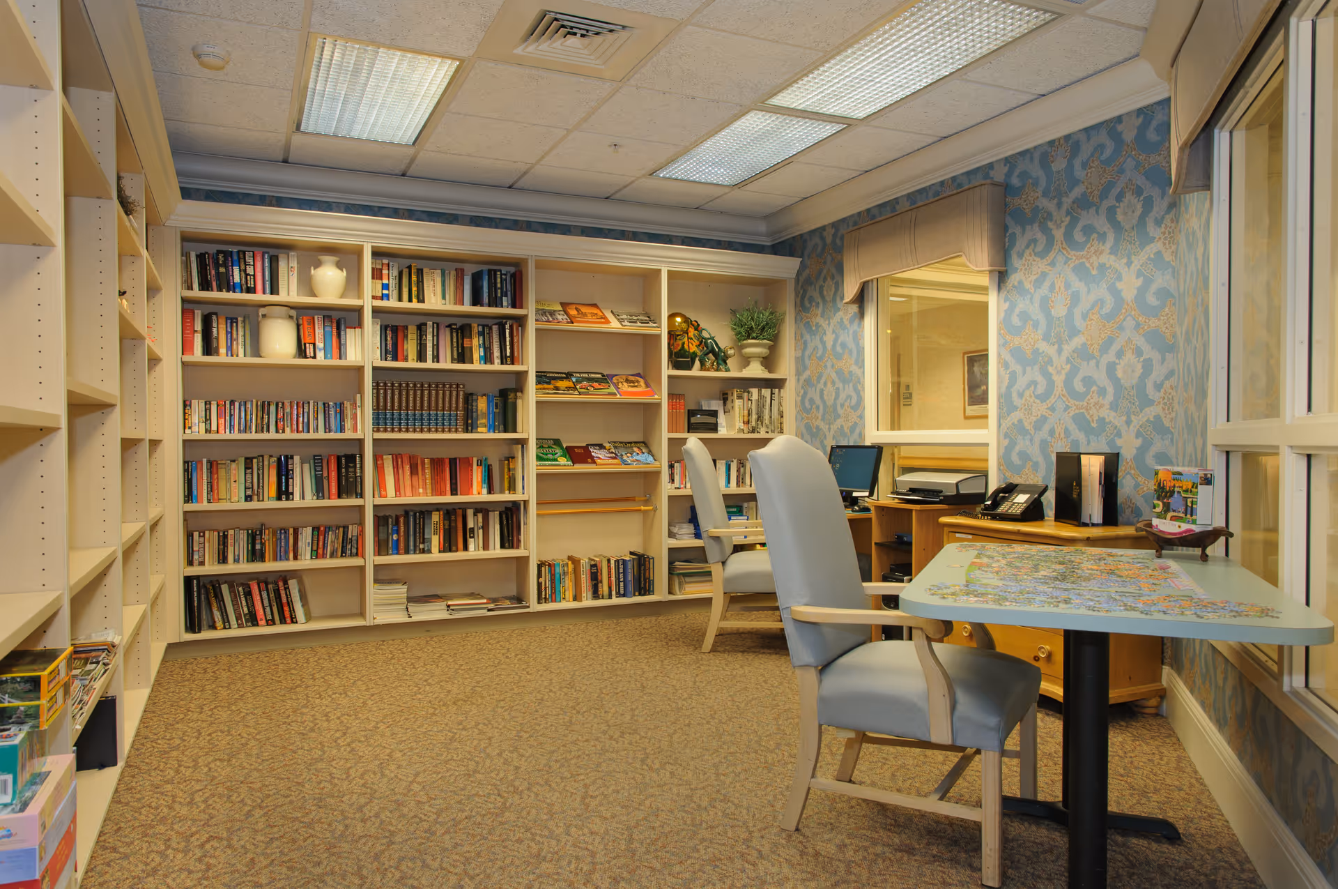A cozy room with bookshelves filled with books and decorative items along the walls, two light blue armchairs, a table with a puzzle in progress, a computer desk with a monitor and printer, and patterned blue wallpaper with a window looking into another room.