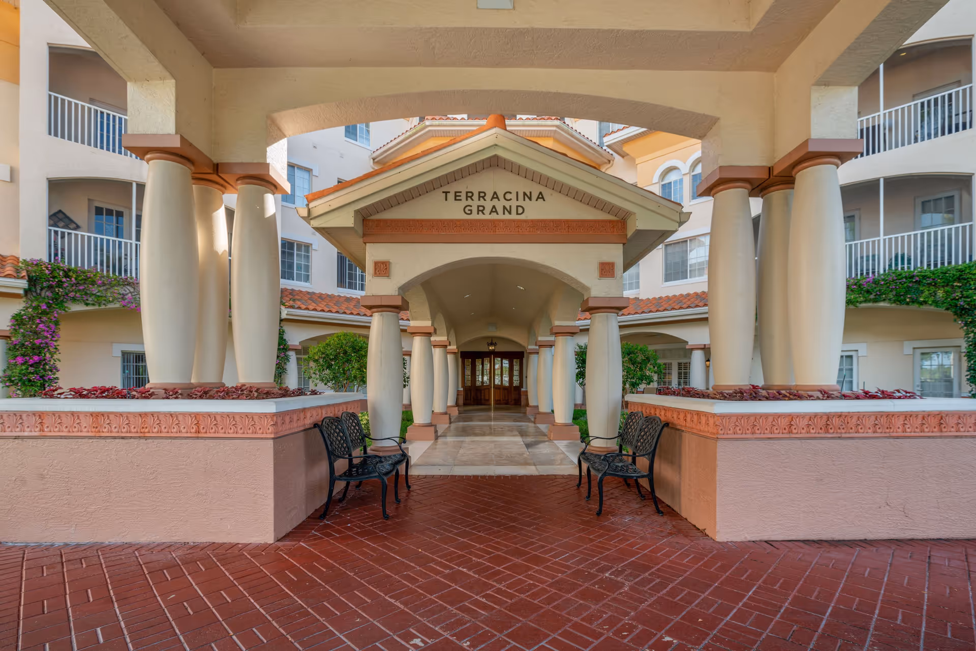 Entrance of Terracina Grand senior living facility featuring a covered walkway with large columns, benches on either side, and a tiled floor leading to glass doors. The building has multiple balconies and is adorned with greenery and flowers.