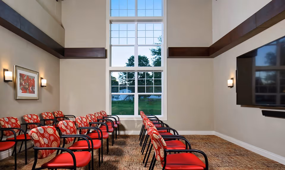 Meeting room with rows of red patterned chairs facing a wall-mounted TV and a tall window.