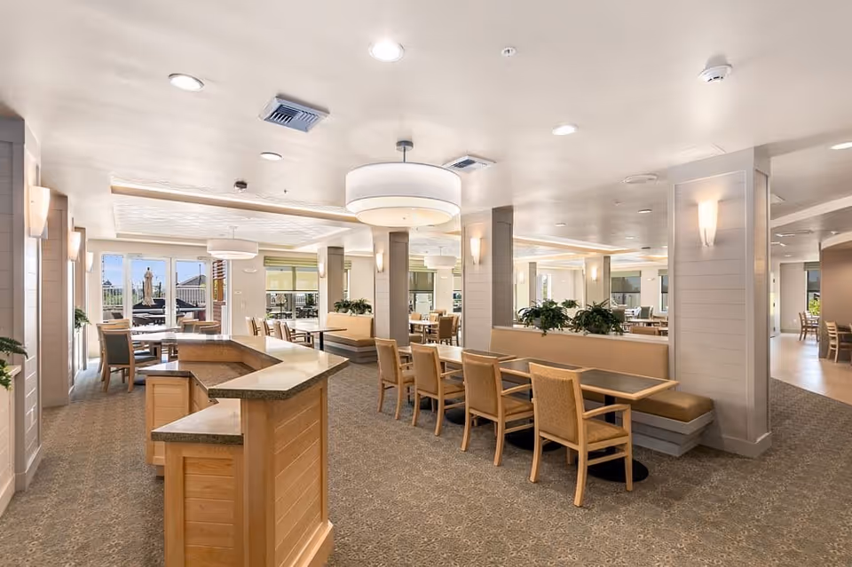 A spacious and well-lit dining area in a senior living facility with multiple tables and chairs arranged neatly. The room features beige walls, carpeted floors, modern light fixtures, and large windows allowing natural light to enter. There are plants placed on dividers between seating areas, and a counter or serving area is visible in the foreground.