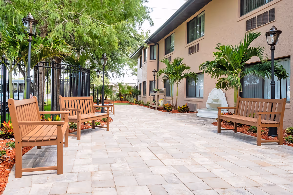 Outdoor patio area at Grand Villa of Englewood featuring a paved walkway with wooden benches on both sides, surrounded by palm trees, plants, and black lamp posts next to a beige building.