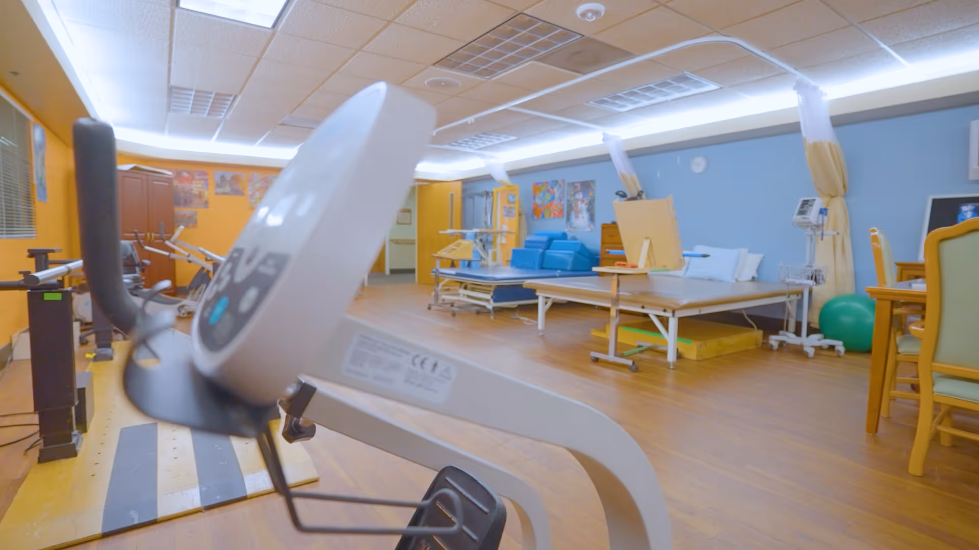 A rehabilitation or physical therapy room with exercise equipment including a stationary bike in the foreground, therapy beds with pillows, a green exercise ball, and chairs. The walls are painted yellow and blue with colorful artwork displayed.