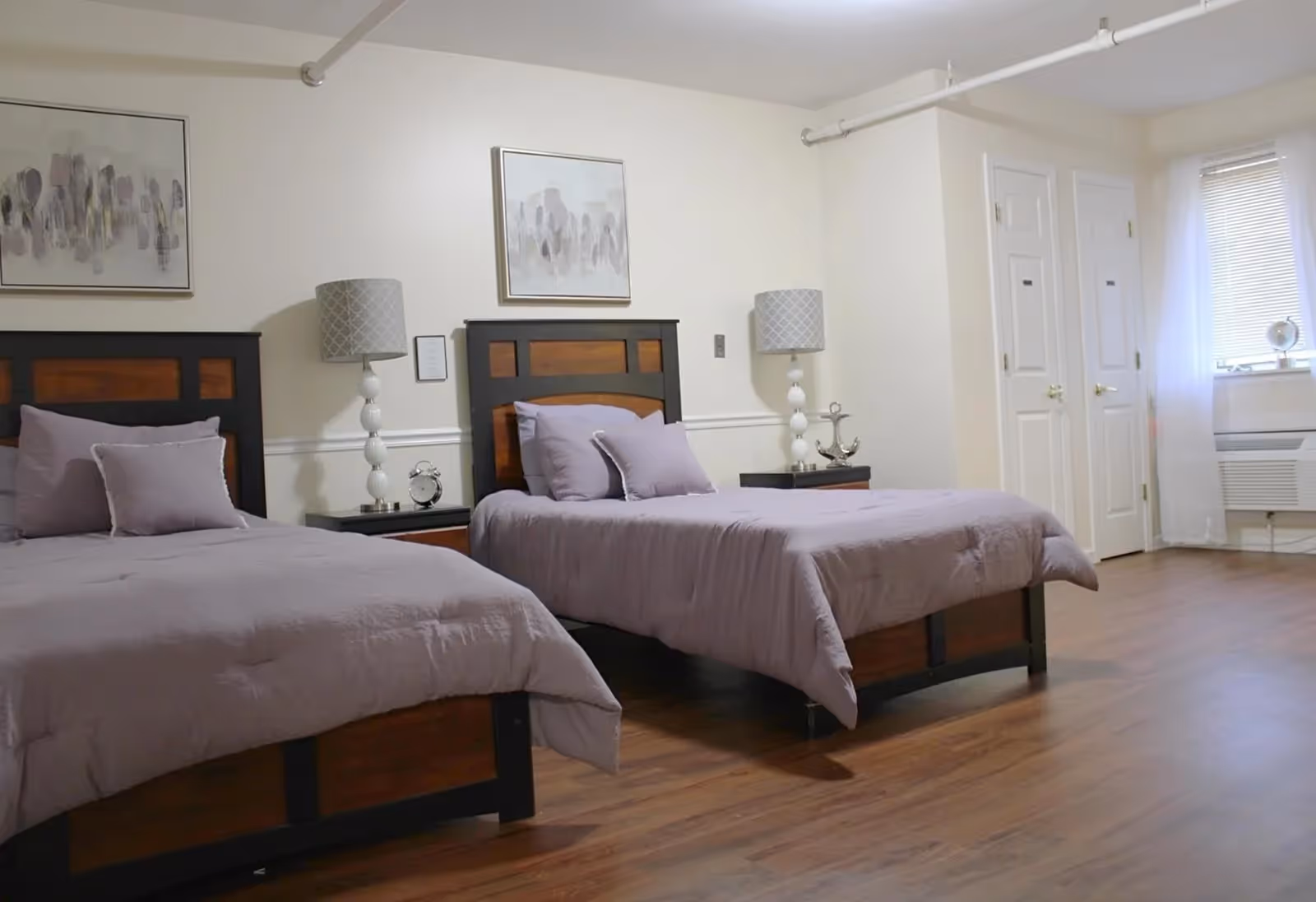 A spacious bedroom with two twin beds featuring wooden headboards and light purple bedding. Each bed has matching pillows and is flanked by a nightstand with a decorative lamp. The room has light-colored walls adorned with abstract artwork, a window with blinds and sheer curtains, and two closed white doors. The floor is wooden.