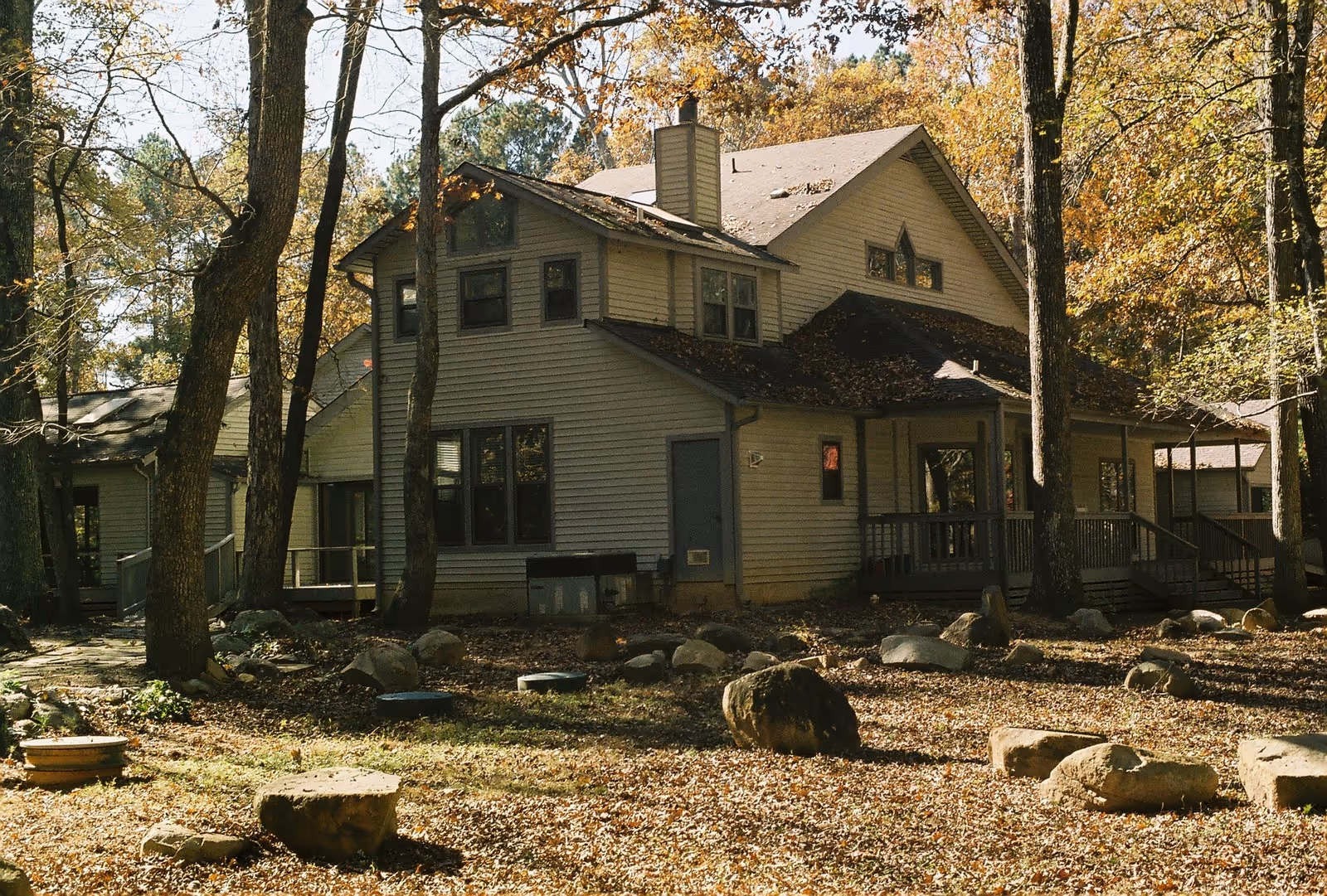 Exterior view of a two-story house surrounded by trees with autumn foliage. The house has beige siding, multiple windows, a chimney, and a covered porch. The ground is covered with fallen leaves and scattered rocks.