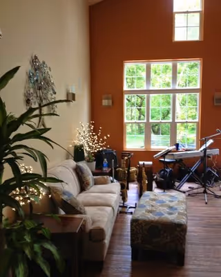Sunlit living room with a sofa, patterned ottoman, large window, plants, and musical equipment.