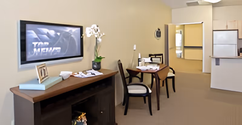 Interior view of a senior living facility room with a small dining table and two chairs, a sideboard with decorative items including a framed photo and a potted orchid, a wall-mounted TV, and a partial view of a kitchen area with a refrigerator and cabinets.