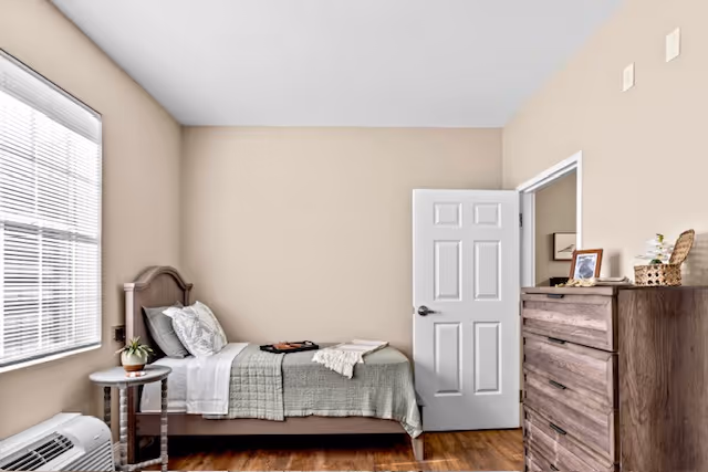 Simple furnished bedroom with a single bed, nightstand, dresser, and a window letting in natural light.