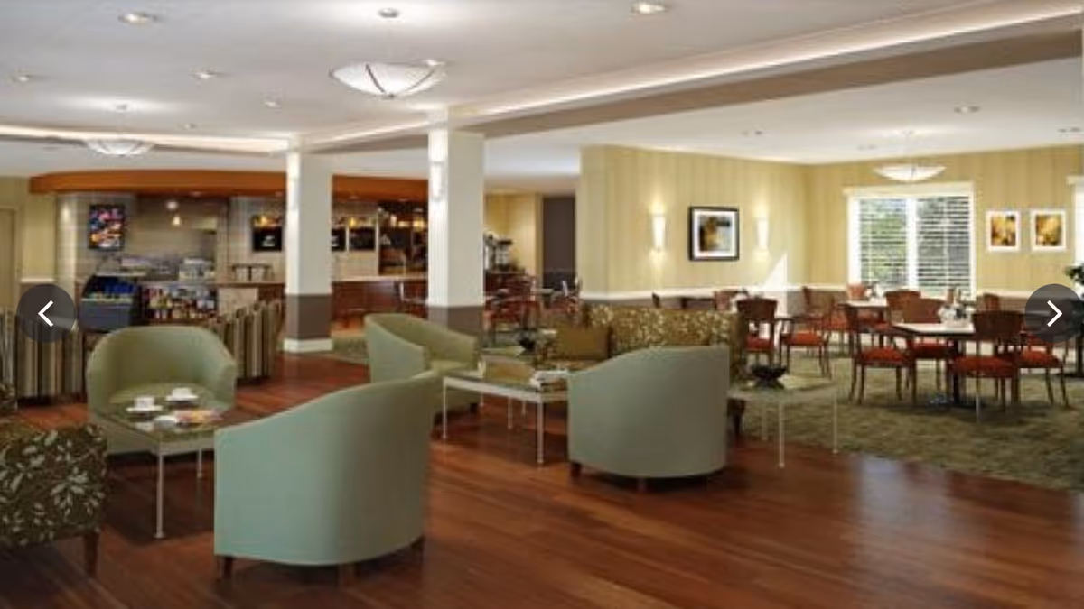 A spacious senior living facility common area with comfortable seating including green armchairs and patterned sofas, wooden flooring, and a dining area with tables and chairs in the background. The room is well-lit with ceiling lights and wall sconces, and there are framed pictures on the walls.