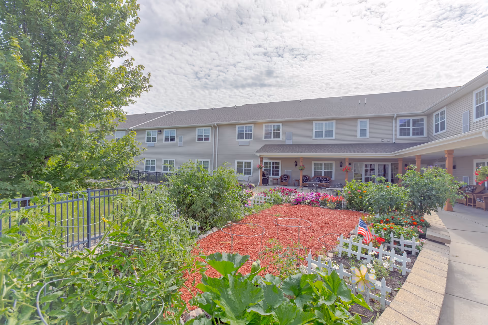 A garden area with various plants and flowers in front of a two-story senior living facility building. The garden includes tomato cages, red mulch, and small white picket fences. The building has beige siding, multiple windows, and a covered patio with seating and hanging flower pots. A large tree is visible on the left side under a partly cloudy sky.