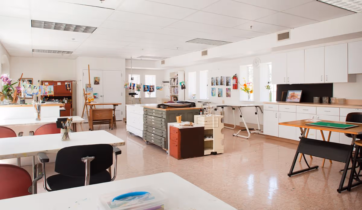 A bright and spacious art studio with white walls and ceiling, several tables and chairs, art supplies like paintbrushes and easels, colorful artwork displayed on the walls and hanging from strings, and cabinets along one side of the room.