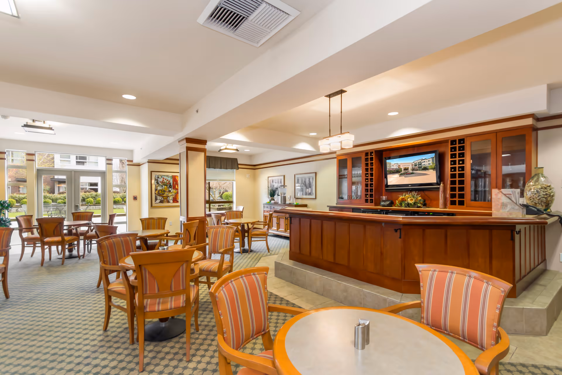 A spacious and well-lit common area in a senior living facility featuring multiple round tables with wooden chairs upholstered in striped fabric. There is a large wooden bar or counter with a mounted flat-screen TV above it, displaying an image of the facility exterior. Large windows and glass doors in the background provide a view of an outdoor garden area with greenery and seating.