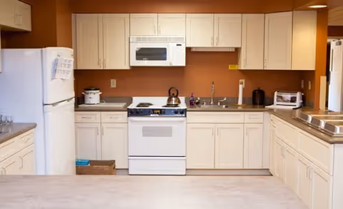 Clean communal kitchen with white cabinets, refrigerator, stove, microwave, and countertops against a warm tan wall.