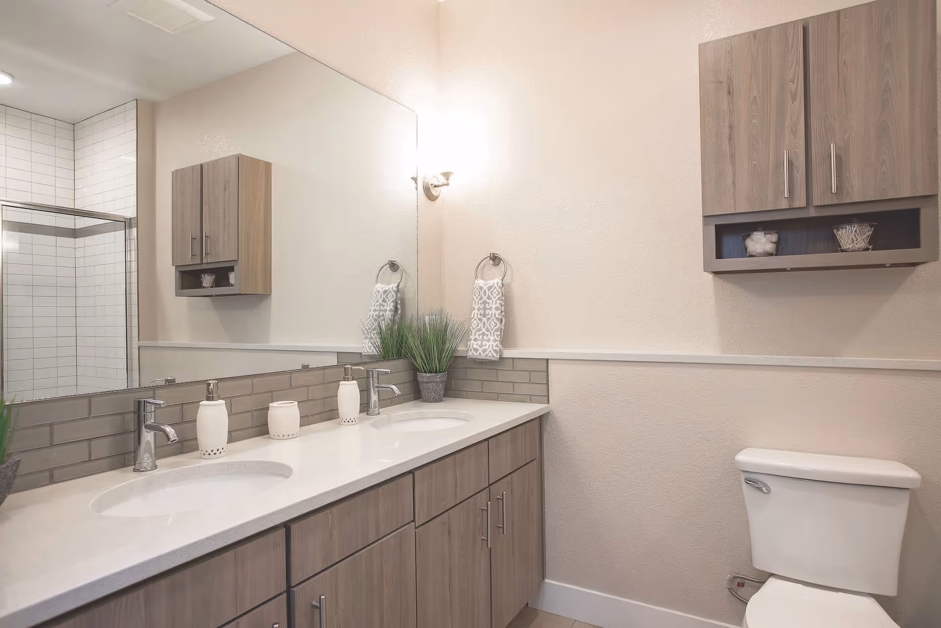 A modern bathroom featuring a double sink vanity with light wood cabinets, two chrome faucets, and white countertop accessories. Above the sinks is a large mirror with a wall-mounted light fixture. To the right, there is a white toilet beneath a matching wooden wall cabinet. In the background, a glass-enclosed shower with white subway tiles is visible.