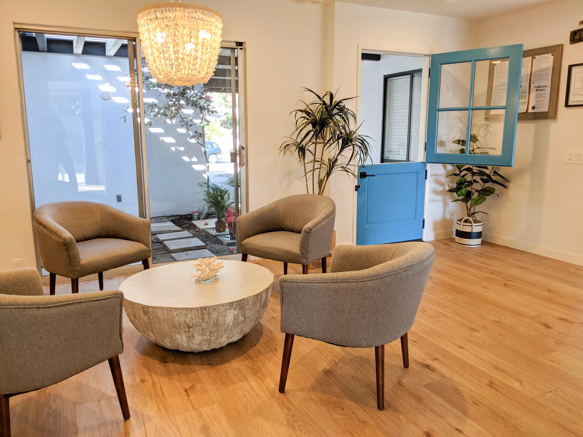 Bright seating area with four gray armchairs arranged around a round stone coffee table, wood floors, plants, a chandelier, and a blue Dutch-style door.