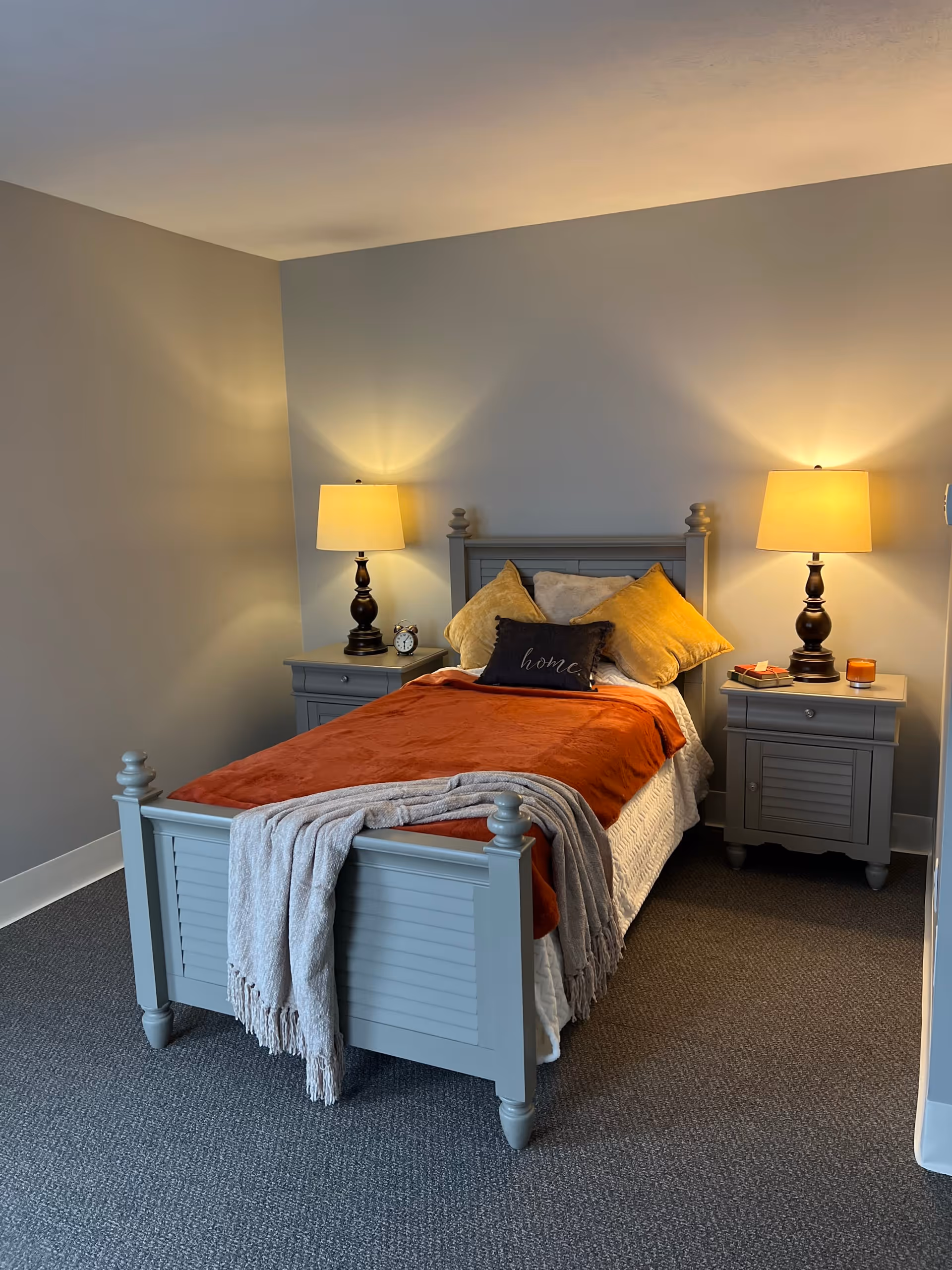 A cozy bedroom with a single gray wooden bed frame, an orange blanket, and multiple pillows including one with the word 'home' embroidered on it. There are two matching gray nightstands on either side of the bed, each with a lit table lamp. One nightstand has a small alarm clock and the other has a candle and a small stack of books. The room has gray carpet and light gray walls.