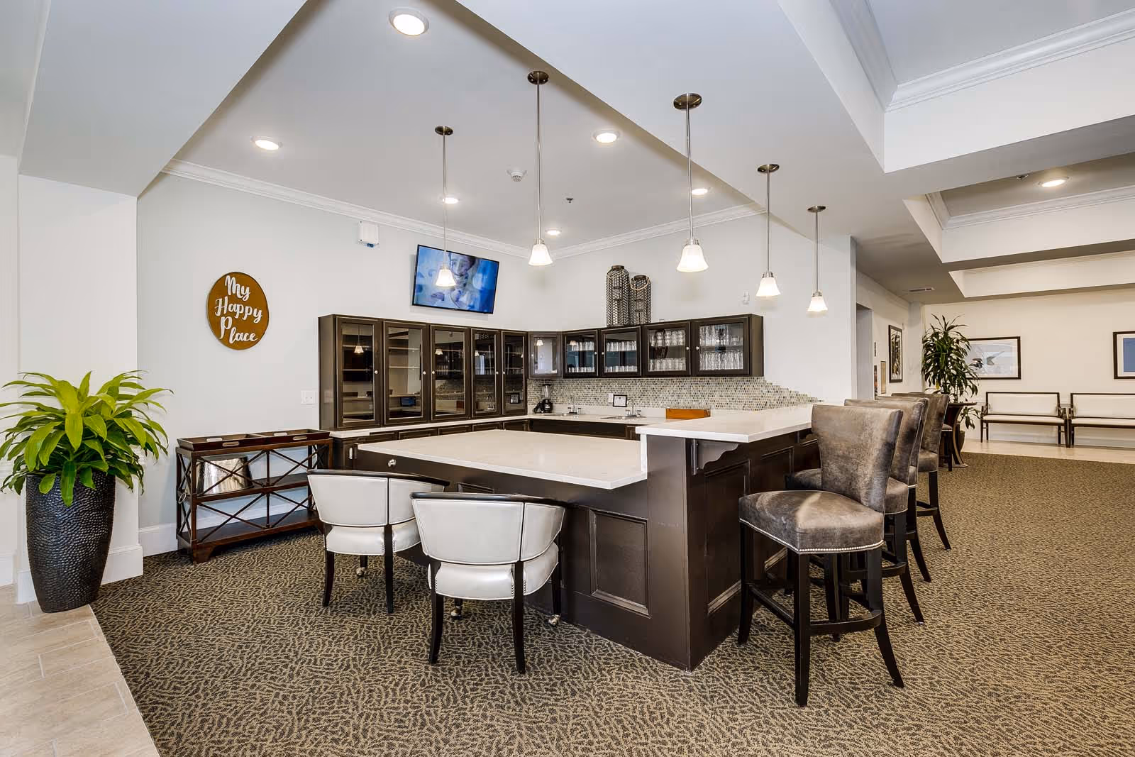 Interior view of a senior living facility lounge area featuring a large dark wood bar with white countertops surrounded by several chairs. There are pendant lights hanging above the bar, a wall-mounted TV, glass-front cabinets, and a decorative sign on the wall that reads 'My Happy Place'. A large potted plant is placed on the left side, and the room has a carpeted floor with neutral tones.