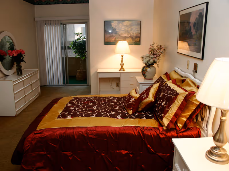 A bedroom with a bed covered in a red and gold patterned comforter and matching pillows. There are two bedside tables with lamps, a dresser with a round mirror and flowers, a desk with a lamp and floral arrangement, and framed artwork on the walls. A sliding glass door with vertical blinds leads to an outdoor area with a potted plant.