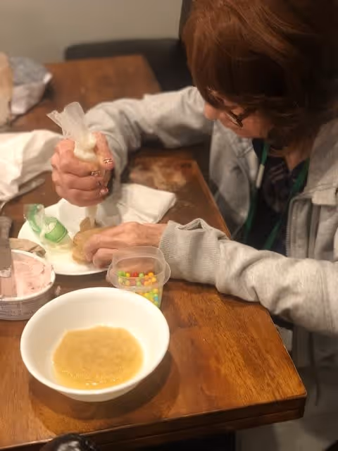 A person with glasses and a gray sweatshirt is sitting at a wooden table, using a piping bag to decorate a cookie or pastry. On the table, there are small containers with colorful candy and other decorating items, as well as a bowl with a light brown mixture.