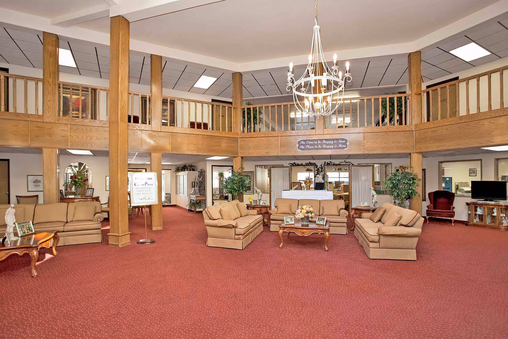 Spacious senior living facility common area with red carpet, beige sofas arranged around wooden coffee tables, wooden pillars and railings on the upper level, a chandelier hanging from the ceiling, and various plants and decorations throughout the room.