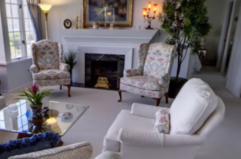 Sunlit living room with a fireplace, floral upholstered armchairs, a white sofa, glass coffee table and potted plants.